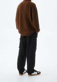 Hnědý žebrovaný svetr, volné černé cargo kalhoty s bílým šitím a černé tenisky s bílými detaily a gumovou podrážkou.