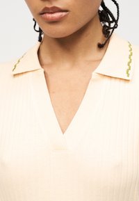 Κοντινό πλάνο ενός ατόμου που φοράει μπλούζα σε κρεμ απόχρωση με ριμπ ύφανση, V λαιμόκοψη και μυτερό γιακά με πράσινες κεντημένες λεπτομέρειες.