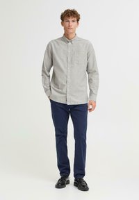 Lys grå skjorte med knapper og en brystlomme, kombinert med marineblå bukser. Modellen har på seg svarte sko og står mot en ensfarget bakgrunn.