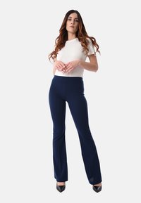 Donna con lunghi capelli mossi, che indossa un top bianco aderente a maniche corte, pantaloni svasati blu navy e tacchi a punta neri, in piedi con le mani Chiuse.
