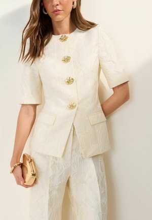 Nő texturált krémszínű, rövid ujjú blézerben aranyszínű virágmintás gombokkal, hozzá illő nadrágban, kezében kis, fényes arany borítéktáskával.