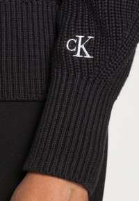 Czarny sweter dziany o prążkowanej fakturze. Na mankiecie znajduje się biały, haftowany logo "ck". Prosty, dopasowany fason.