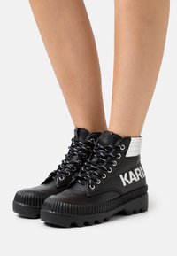 Botas altas de cuero negro con suelas gruesas y estriadas, cordones en blanco y negro, panel blanco en los tobillos y la marca "KARL" en el lateral, llevadas sobre piernas desnudas.