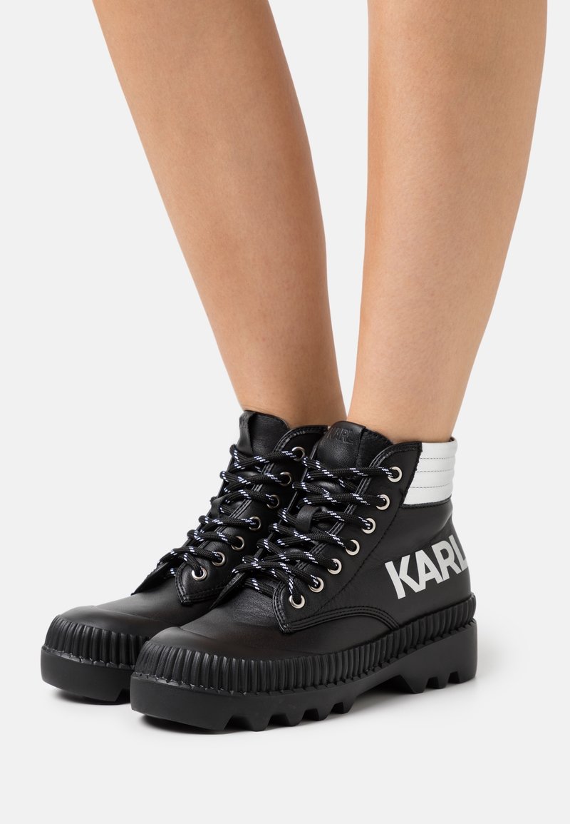 Botas altas de cuero negro con suelas gruesas y estriadas, cordones en blanco y negro, panel blanco en los tobillos y la marca "KARL" en el lateral, llevadas sobre piernas desnudas.