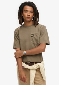 Camiseta de algodón verde oliva con un bolsillo en el pecho, combinada con pantalones beige y un cinturón crema. Acentuada por un reloj y peinada con rastas.