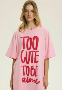Młoda kobieta o blond włosach nosi oversize różową koszulę z wyraźnym czerwonym napisem "TOO CUTE TO BE alone" na jasnym tle.