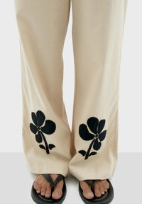 Pantaloni beige a gamba larga con ricami floreali neri ai polsini. Il materiale sembra liscio, abbinato a infradito nere.