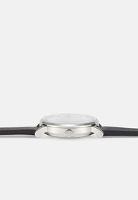 Reloj de acero inoxidable plateado con correa de cuero negra, esfera redonda, cubierta de cristal y una corona lateral para ajustes. Diseño simple y minimalista.