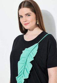 Camiseta negra con un gran aplique de hoja arrugada en verde menta a lo largo del hombro. Tejido suave con un ajuste relajado y escote redondo.