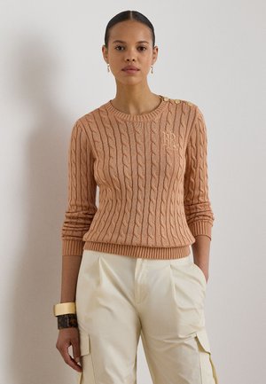 Kvinde iført en ferskenfarvet kabelstrikket sweater med knapdetaljer på skulderen og cremefarvede cargobukser, stående med den ene hånd i lommen.