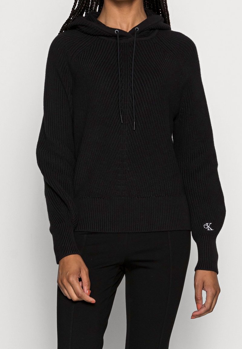 Czarna dzianinowa bluza z kapturem o prążkowanej fakturze, kapturem z troczkiem i logo na mankiecie. Luźny krój, długie rękawy i ściągacz na dole.