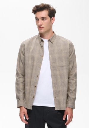 Jeune homme portant une chemise beige boutonnée par-dessus un t-shirt blanc et un pantalon noir, debout devant un fond blanc uni.