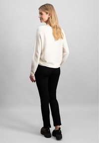 Tejfehér pulóver, eső vállú, fekete, slim-fit nadrággal és masszív fekete cipővel párosítva, hátulról nézve. Az összhatás sima textúrájú.