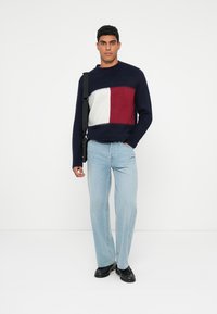Námornícky sveter s veľkým červenobielym geometrickým nápatchom; skombinovaný s svetlými modrými džínsami; slim fit; model drží čiernu tašku na rameno.