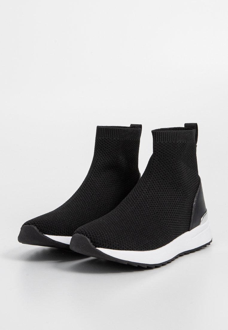 Bottines en tricot noir avec un design de type chaussette, dotées d'une semelle extérieure blanche et d'accents noirs brillants près du talon.