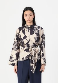 Blusa crema con motivo floreale scuro, con colletto, cintrata in vita, caratterizzata da maniche lunghe e larghe e una texture liscia.