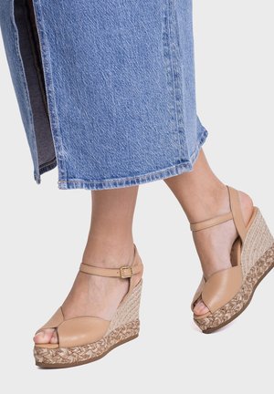 Pies con sandalias de cuña beige con tiras en el tobillo y vaqueros azules de corte ancho y cropped sobre fondo claro.