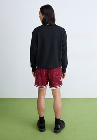 Schwarzer Sweatshirt, rote Shorts mit marmoriertem Muster und weißen Streifen, schwarze Socken und schwarze Schuhe, stehend auf grünem Boden.