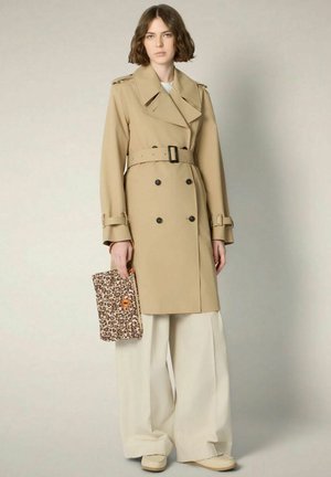 Donna con trench beige doppiopetto, pantaloni bianchi larghi, scarpe crema, che tiene una pochette con stampa leopardata.