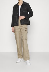 Osoba oblečená v černé bundě s bílým prošíváním, béžových cargo kalhotách s bočními kapsami a bílých teniskách, stojící na jednorázovém bílém pozadí.