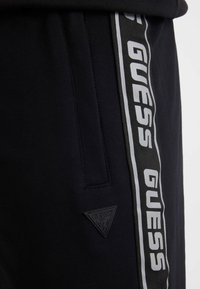 Pantalones de chándal negros con una franja lateral que lleva "GUESS" en blanco y gris, tejido texturizado y un parche con un logo triangular.