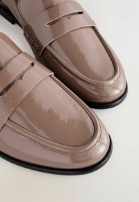 Glänzende, hellbraune Lacklederslipper mit einem Band über der Oberseite, abgerundeter Zehenpartie und schwarzer Gummisohle. Strukturiertes Nähen ist sichtbar.