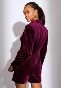 Burgundi bársony zakó strukturált vállakkal és passzoló sorttal. Jellemzője a fényes textura és a gombos díszítés.