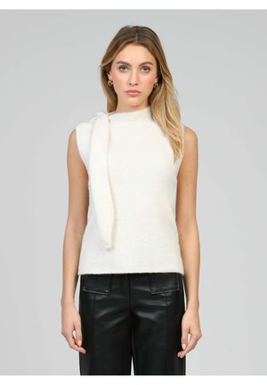 Femme portant un haut en maille blanc sans manches avec un détail noué sur une épaule et un pantalon en cuir noir, debout devant un fond gris uni.