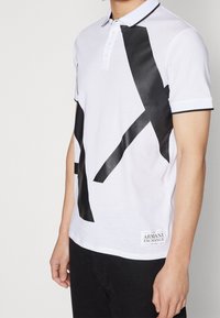 Biała koszulka polo z czarnym abstrakcyjnym wzorem, krótki rękaw, zapięcie na guziki z kołnierzykiem, kontrastowe wykończenia i metka marki na dole.
