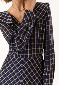 Kobieta w granatowej sukience w beżową kratkę, z długimi rękawami i srebrnym naszyjnikiem.