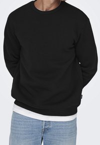 Felpa nera con collo a girocollo in misto cotone, con colletto e polsini a coste, indossata sopra una t-shirt bianca, abbinata a jeans blu.