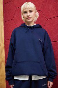Felpa con cappuccio blu navy in tessuto morbido, con tasca a marsupio, che presenta un piccolo logo bianco stampato sul petto, indossata sopra una maglietta bianca.