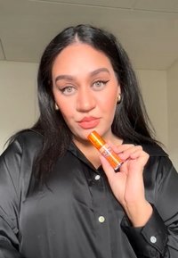 Produit cosmétique orange avec un capuchon transparent, tenu par une personne portant une chemise en satin noir. De longs cheveux noirs et un maquillage subtil sont visibles.