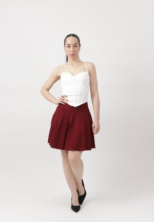 Kobieta stojąca na białym tle, ubrana w biały bezrękawnik, czerwoną plisowaną spódnicę i czarne szpilki, z ręką na biodrze i neutralnym wyrazem twarzy.
