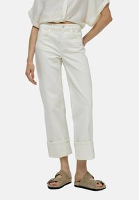 Jeans de mezclilla blanca recortados con puños enrollados, que cuentan con un cierre de botón y bolsillos frontales. Combinados con sandalias marrones tipo slip-on.