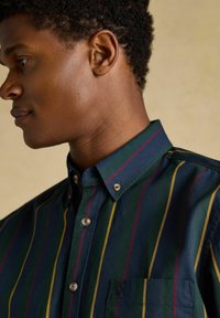 Mladý muž v košeli s pruhmi v zelenej, námorníckej, žltej a červenej farbe, pozerajúci doľava na pozadí s jednofarebným béžovým pozadím.