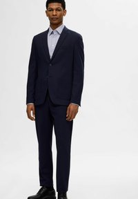 Abito blu navy con giacca a due bottoni e tasche applicate, abbinato a una camicia a righe. Il tessuto ha una texture liscia, completato da scarpe nere.