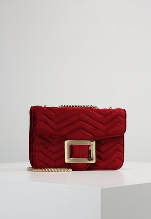 Petit sac à main en velours rouge avec surpiqûres en chevrons, boucle rectangulaire dorée et bandoulière chaîne dorée sur une surface blanche avec un fond gris.
