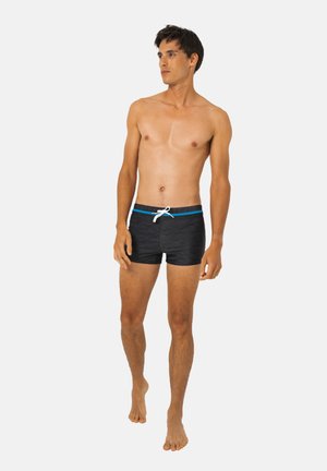 Jeune homme aux cheveux courts et foncés, portant un short de bain noir avec une bordure bleue et un cordon blanc, debout pieds nus devant un fond blanc.