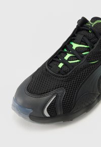Czarne sportowe buty z siateczkową cholewką, zielonymi sznurówkami i gumowymi akcentami. Posiadają zaokrąglony czubek oraz chropowate boki dla lepszej przyczepności i wsparcia.