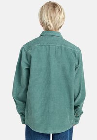 Camicia in velluto a coste di un blu verde chiaro, con colletto classico, maniche lunghe e polsini abbottonati; texture a coste con orlo leggermente curvo sul retro.