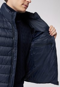 Marineblaue, gepolsterte Jacke mit Reißverschluss vorne, die ein gestepptes Muster aufweist und auf der linken Seite eine Innentasche mit Reißverschluss hat.