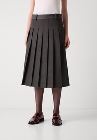 Personne portant une jupe midi plissée gris foncé, des collants noirs transparents, des mocassins bordeaux avec des détails dorés, et un haut blanc, se tenant sur un sol blanc.