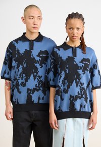 Πλεκτά πόλο μπλουζάκια σε μπλε χρώμα με μαύρο αφηρημένο μοτίβο, με μαύρο γιακά και τριπλό κουμπί, φορεμένα από δύο μοντέλα.