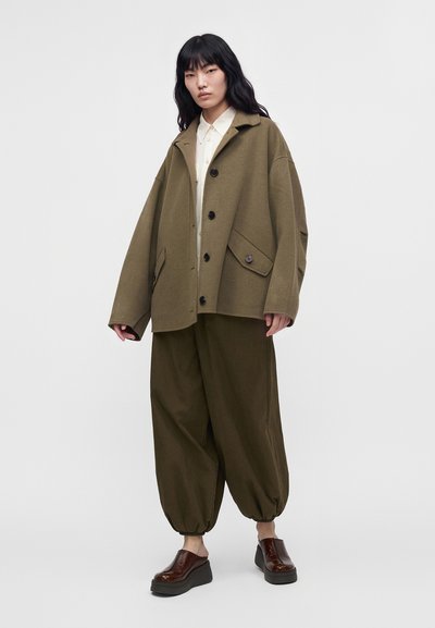 Mujer con pelo largo y negro que lleva una chaqueta holgada verde oliva, pantalones sueltos de color oliva con puños elásticos, camisa blanca y zapatos plataforma oscuros.