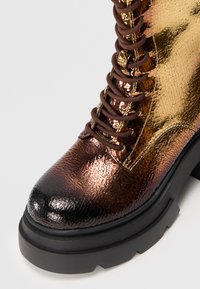 Bottine à lacets avec une finition métallique craquelée dans des teintes de bronze et d'or, présentant un bout rond et une semelle épaisse et robuste en noir.
