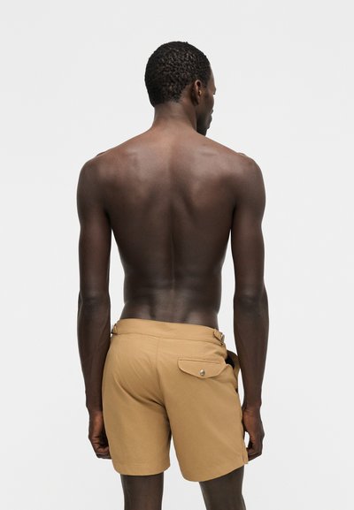 Rövid, göndör hajú férfi, ing nélkül, bézs rövidnadrágot visel hátsó zsebbel, oldalra fordulva egy egyszerű világos háttér előtt áll.