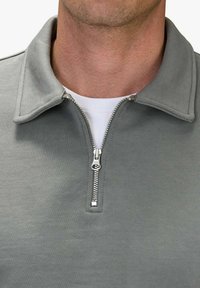 Grå zip-up krage sweatshirt med en slät textur, med en silverfärgad metalldragkedja och en vit undertröja som syns under.