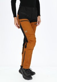 Pantaloni marrone-arancio e neri, caratterizzati da numerose tasche con zip, un tessuto testurizzato e una vita regolabile. Decorati con un patch del logo.