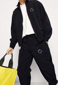 Chándal deportivo negro hecho de material ligero, con chaqueta con cremallera y pantalones con cordón, ambos con detalle de logo. Bolsa de mano amarilla.